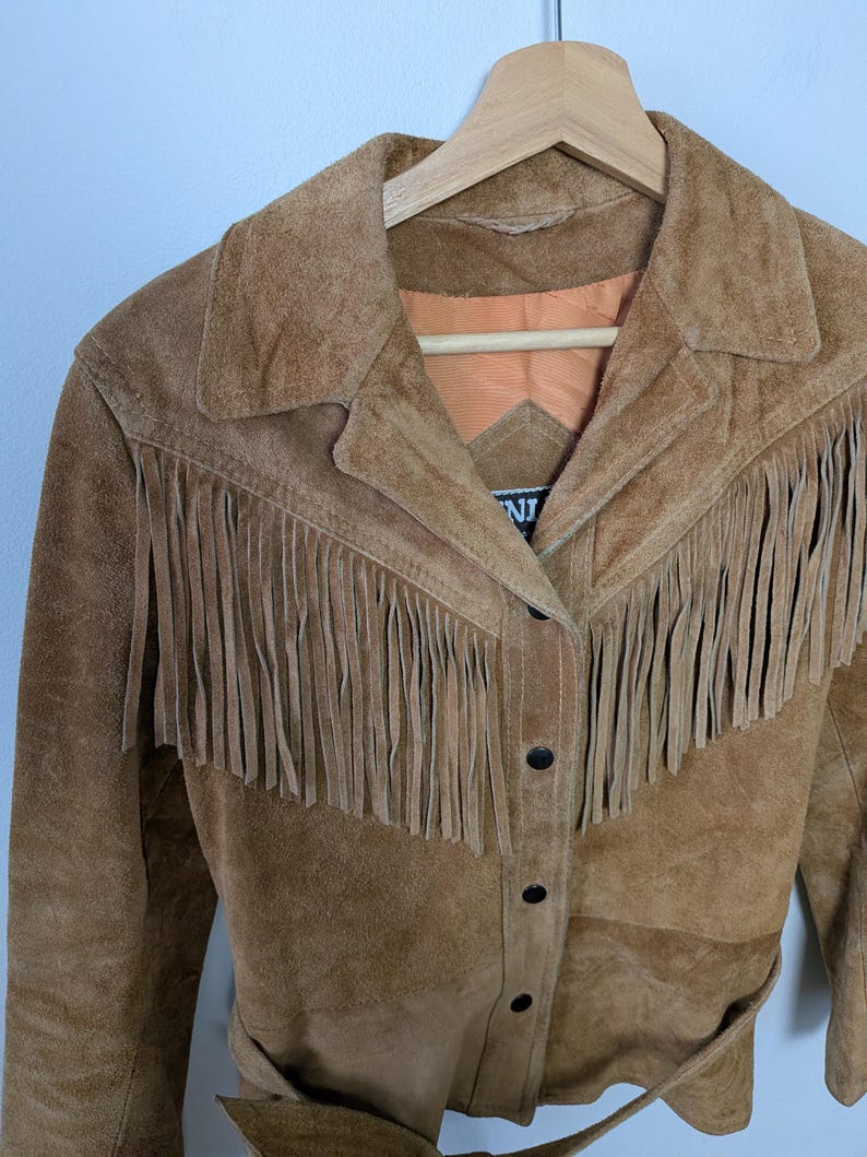 May include: A tan suede jacket featuring long fringe detailing across the chest and shoulders. The jacket has a classic collar, a button-front closure with black buttons, and an orange interior lining. A partial brand label is visible at the neck.