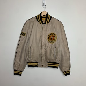 May include: A beige bomber jacket with black and yellow striped ribbing. The jacket has a zipper closure and a large embroidered patch on the left side that reads "Steel Workers of America, AFL-CIO, Local 3589".