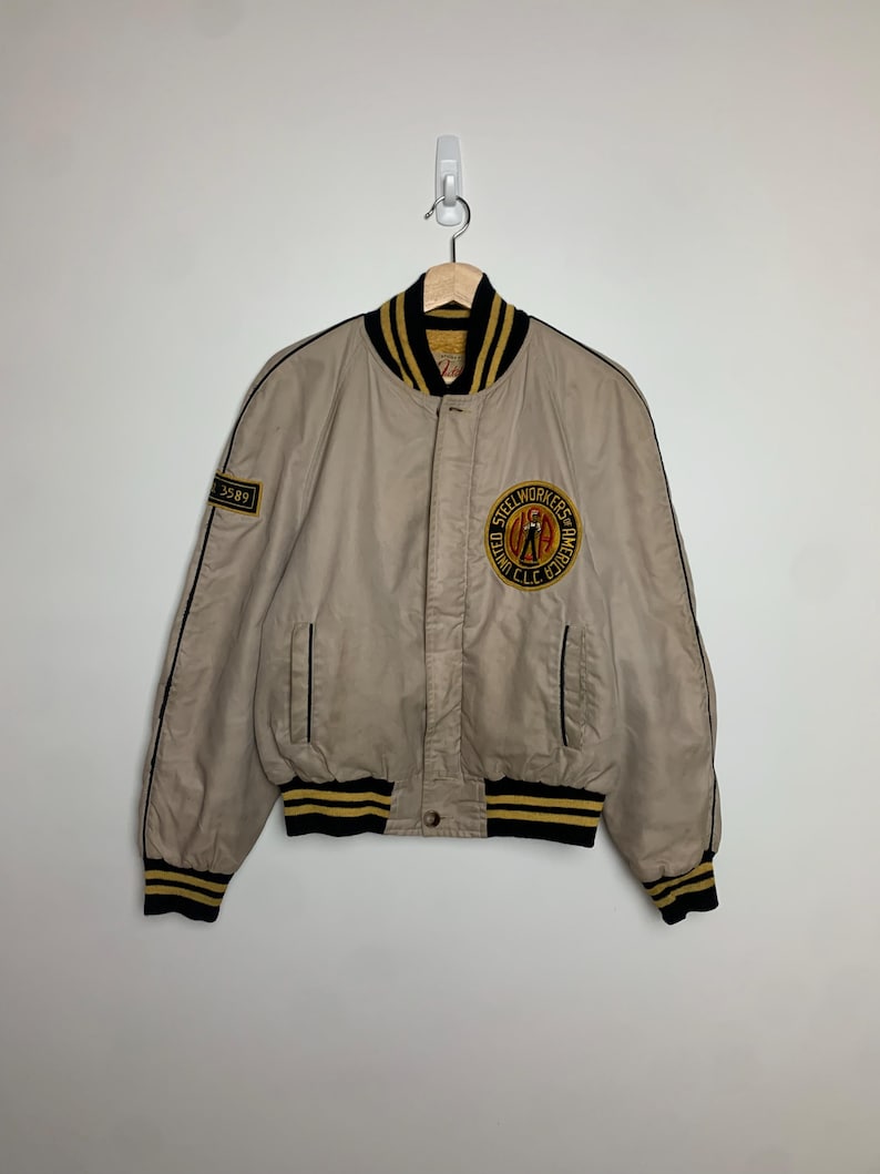May include: A beige bomber jacket with black and yellow striped ribbing. The jacket has a zipper closure and a large embroidered patch on the left side that reads "Steel Workers of America, AFL-CIO, Local 3589".