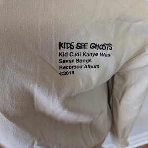 May include: A beige long-sleeve sweatshirt featuring black printed text that reads KIDS SEE GHOSTS, Kid Cudi Kanye West, Seven Songs, Recorded Album, and &copy;2018. The garment has a plain, solid-colored fabric surface with a simple crew neckline.