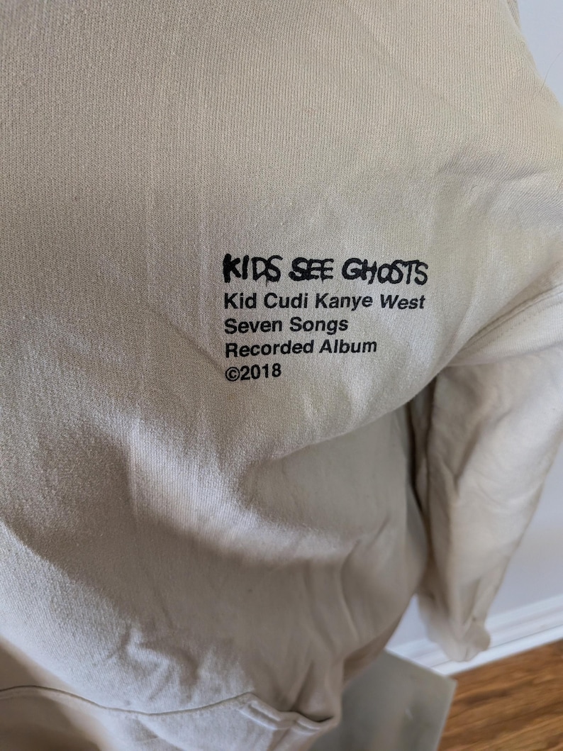 May include: A beige long-sleeve sweatshirt featuring black printed text that reads KIDS SEE GHOSTS, Kid Cudi Kanye West, Seven Songs, Recorded Album, and &copy;2018. The garment has a plain, solid-colored fabric surface with a simple crew neckline.