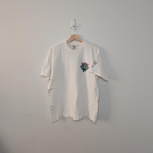May include: A white short-sleeve t-shirt featuring a small graphic on the left chest depicting a baseball diamond with the text "The Florida Marlins" and a small logo on the left sleeve. The shirt is displayed on a wooden hanger against a plain wall.