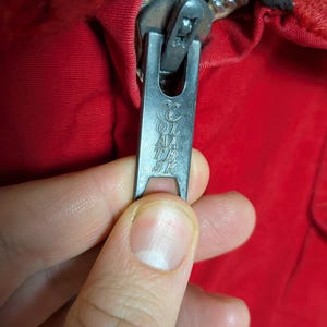 May include: Close-up of a silver zipper pull with the text "COL AA TR SK" on a red garment. The zipper is partially open, revealing the teeth and the fabric's texture. The zipper pull is held by a hand.