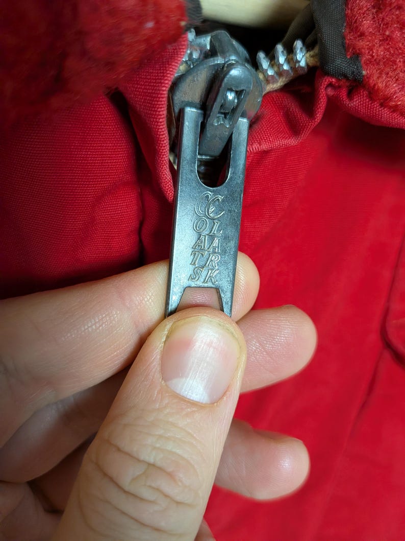 May include: Close-up of a silver zipper pull with the text "COL AA TR SK" on a red garment. The zipper is partially open, revealing the teeth and the fabric's texture. The zipper pull is held by a hand.