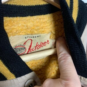 May include: Close-up of a beige jacket with a yellow and black striped collar. The jacket has a red label that says "Styled by Outdoor" and a smaller label that says "Our Emblem".