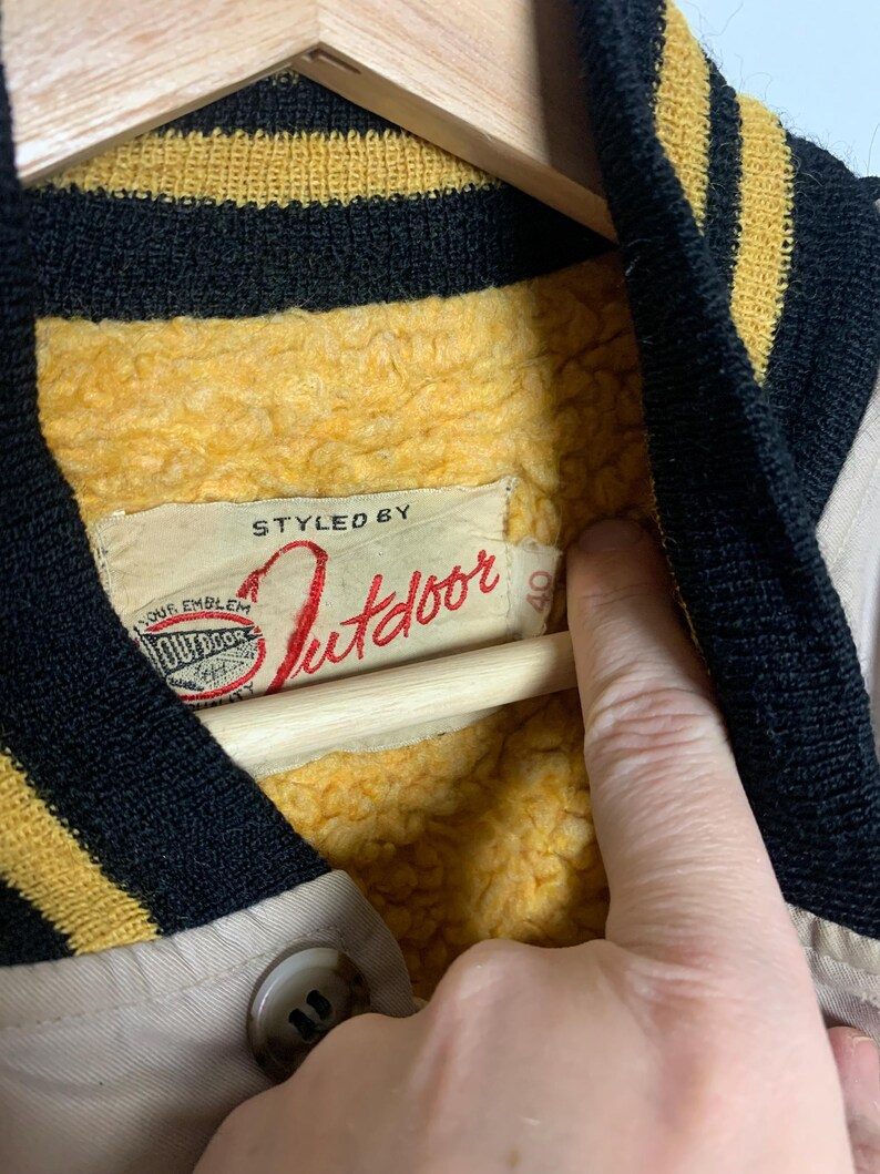 May include: Close-up of a beige jacket with a yellow and black striped collar. The jacket has a red label that says "Styled by Outdoor" and a smaller label that says "Our Emblem".