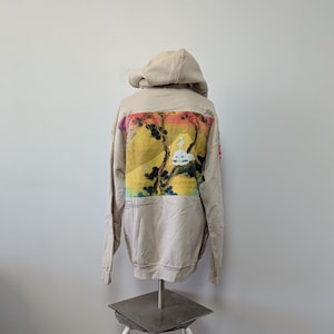 May include: A beige hooded sweatshirt featuring a large rectangular graphic on the back. The print displays a yellow and orange landscape with dark tree silhouettes and a small white figure on a cloud. Red text is visible on the right sleeve.