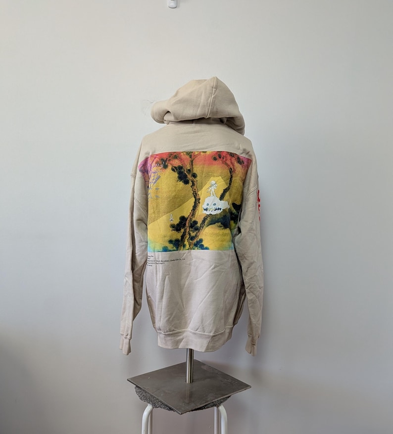 May include: A beige hooded sweatshirt featuring a large rectangular graphic on the back. The print displays a yellow and orange landscape with dark tree silhouettes and a small white figure on a cloud. Red text is visible on the right sleeve.