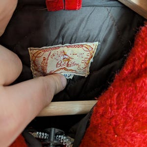 May include: Close-up of a vintage red jacket with a zipper and a label. The label reads "Original Bob Allen" with a fishing-themed illustration. The jacket has a quilted lining and a size 42 tag.
