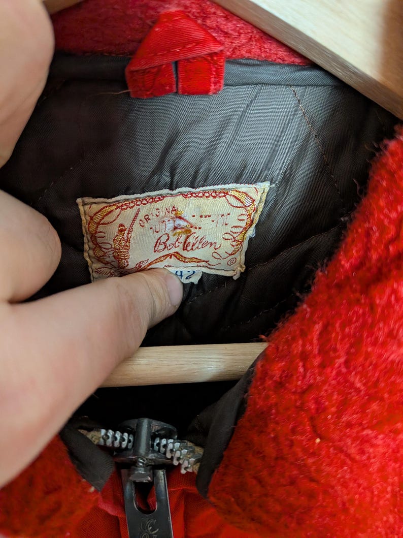 May include: Close-up of a vintage red jacket with a zipper and a label. The label reads "Original Bob Allen" with a fishing-themed illustration. The jacket has a quilted lining and a size 42 tag.