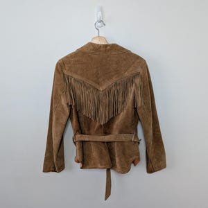 May include: A brown suede jacket featuring long fringe detailing across the upper back section. The jacket includes a matching suede belt tied around the waist, long sleeves, and a pointed collar, all displayed on a wooden hanger against a wall.