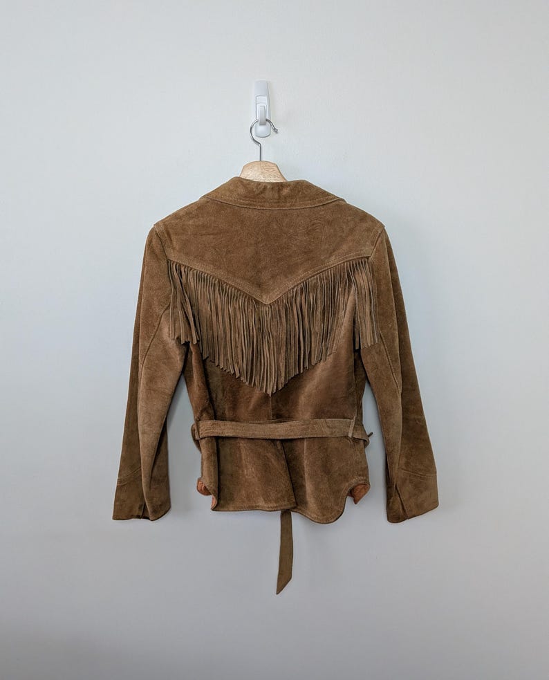 May include: A brown suede jacket featuring long fringe detailing across the upper back section. The jacket includes a matching suede belt tied around the waist, long sleeves, and a pointed collar, all displayed on a wooden hanger against a wall.