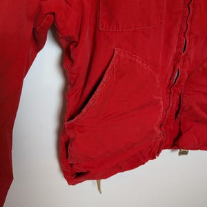 May include: Close-up of a red jacket with a pocket and a zipper. The jacket is made of a soft material and has a worn appearance. The jacket's colour is a vibrant red.