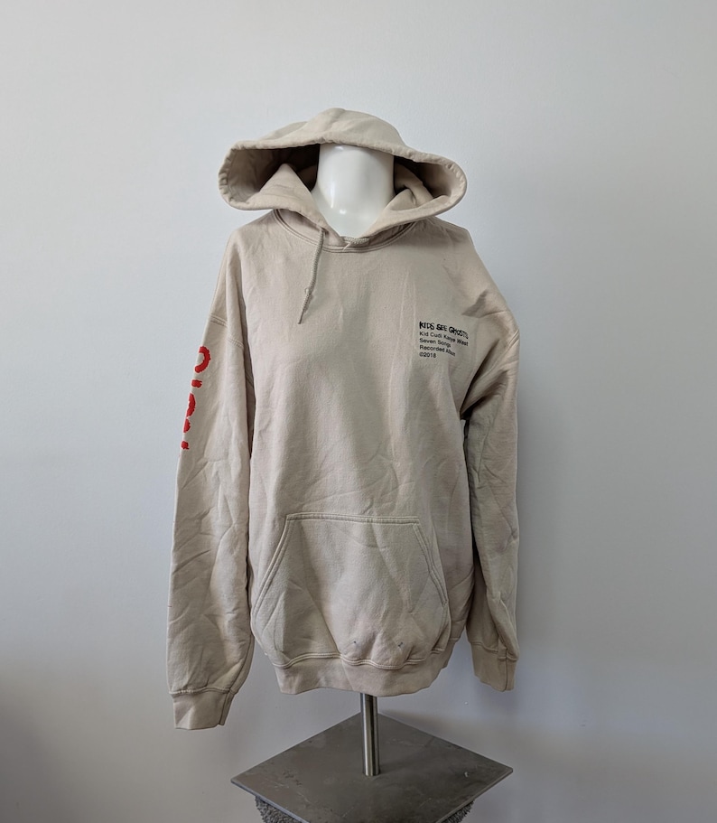 May include: A beige hooded sweatshirt featuring a front kangaroo pocket and long sleeves. The chest displays black text reading Kids See Ghosts, and the right sleeve features red text. The hoodie is displayed on a mannequin torso.