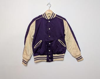 May include: A purple and cream-colored varsity jacket with a button-up front, two front pockets, and striped trim on the cuffs, collar, and hem. The jacket is hanging on a wooden hanger against a white wall.