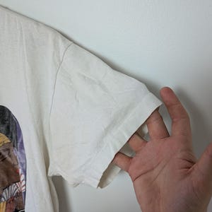 May include: A white t-shirt with a graphic design on the front. The sleeve of the shirt is visible, and a hand is touching the sleeve. The shirt has a vintage look.