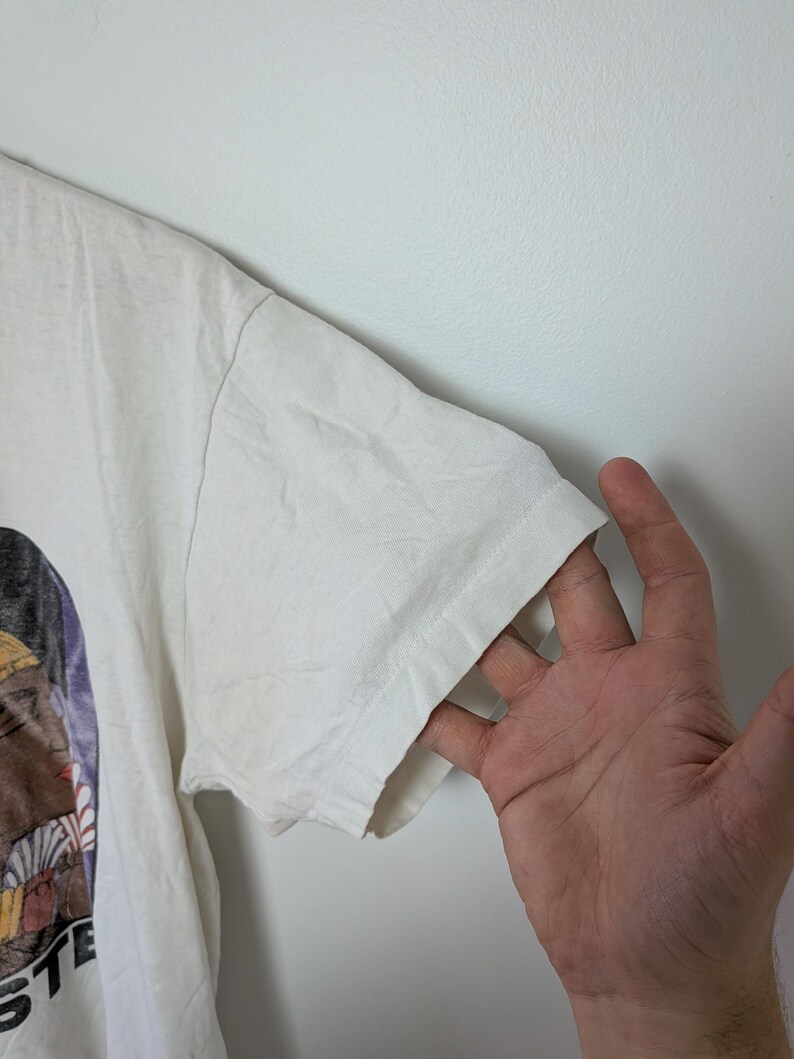 May include: A white t-shirt with a graphic design on the front. The sleeve of the shirt is visible, and a hand is touching the sleeve. The shirt has a vintage look.