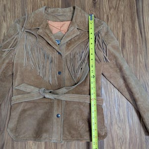 May include: A tan suede jacket featuring long fringe detailing across the chest and shoulders, a tie-waist belt, and a button-front closure. A yellow measuring tape is placed vertically along the front, showing a length of approximately 26 inches.