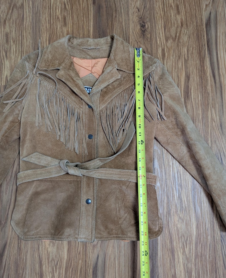 May include: A tan suede jacket featuring long fringe detailing across the chest and shoulders, a tie-waist belt, and a button-front closure. A yellow measuring tape is placed vertically along the front, showing a length of approximately 26 inches.