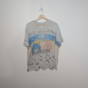 May include: A vintage, off-white t-shirt with a graphic featuring the words "LOVE MACHINE" and "DURACELL". The design includes a blue and pink cartoon image with a landscape background. The shirt is hanging on a wooden hanger.
