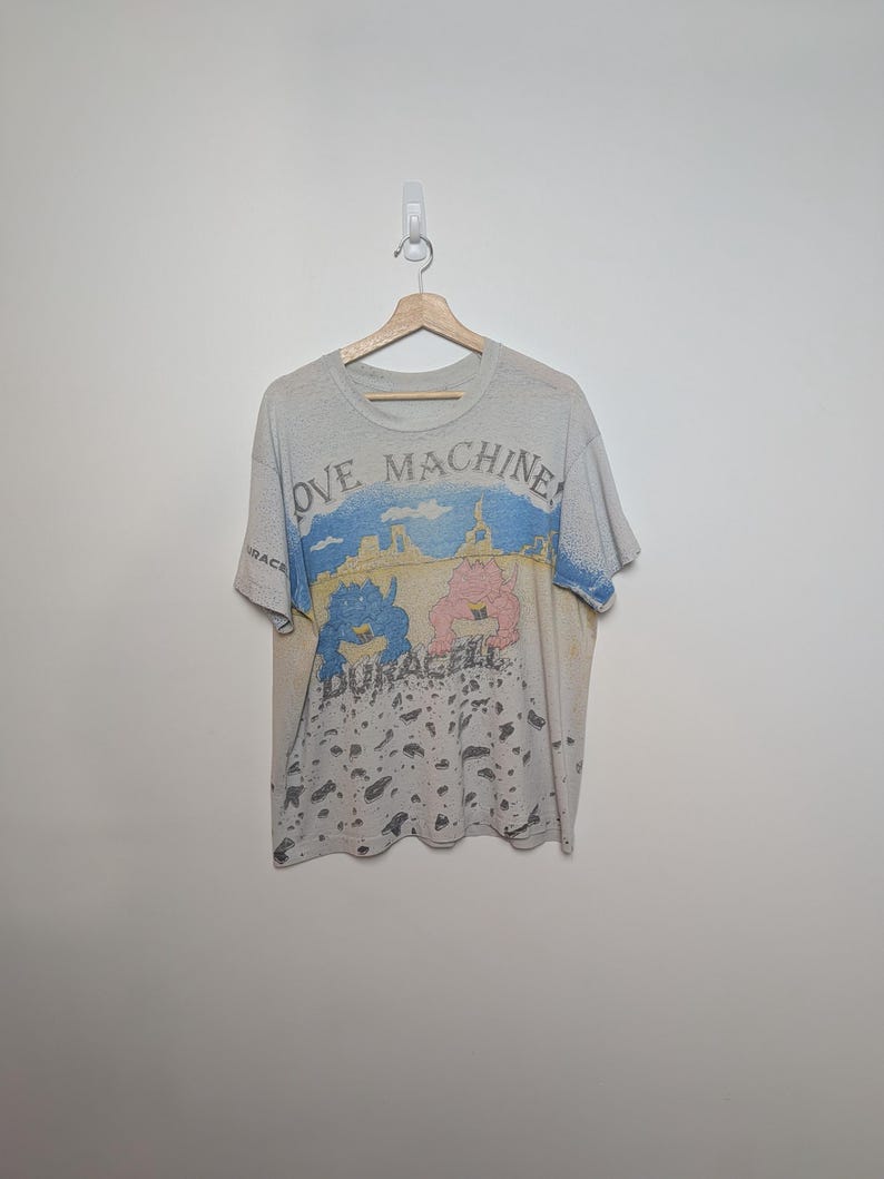 May include: A vintage, off-white t-shirt with a graphic featuring the words "LOVE MACHINE" and "DURACELL". The design includes a blue and pink cartoon image with a landscape background. The shirt is hanging on a wooden hanger.
