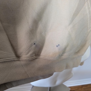 May include: A beige hooded sweatshirt featuring a ribbed hem and two small, dark, circular stains located near the bottom front edge of the fabric. The garment is constructed from a solid-colored knit material with visible stitching along the hemline.