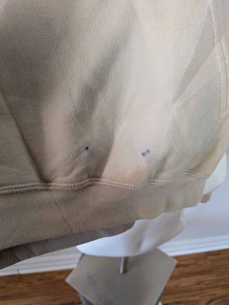 May include: A beige hooded sweatshirt featuring a ribbed hem and two small, dark, circular stains located near the bottom front edge of the fabric. The garment is constructed from a solid-colored knit material with visible stitching along the hemline.