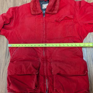 May include: A red corduroy jacket with a faux fur collar. The jacket has a zipper closure, multiple pockets, and a tan strap at the bottom. The jacket is approximately 58 cm long.