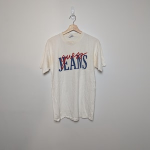 May include: A cream-coloured t-shirt featuring the text "GUESS JEANS" in red and blue. The t-shirt is hanging on a wooden hanger against a plain white wall. The shirt has short sleeves and a crew neck.