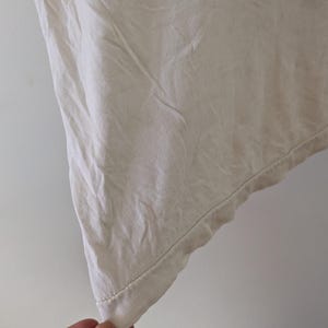 May include: Close-up of a cream-colored fabric, possibly linen or cotton, with visible texture and slight wrinkles. The edge of the fabric is visible, showing a hem with a subtle stitch. The fabric appears to be part of a garment.