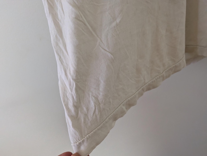 May include: Close-up of a cream-colored fabric, possibly linen or cotton, with visible texture and slight wrinkles. The edge of the fabric is visible, showing a hem with a subtle stitch. The fabric appears to be part of a garment.