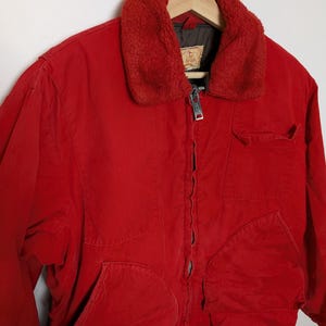 May include: A vibrant red jacket featuring a plush, reddish-brown collar. The jacket has a zip closure, two large front pockets, and a smaller chest pocket. The jacket is suspended on a wooden hanger.