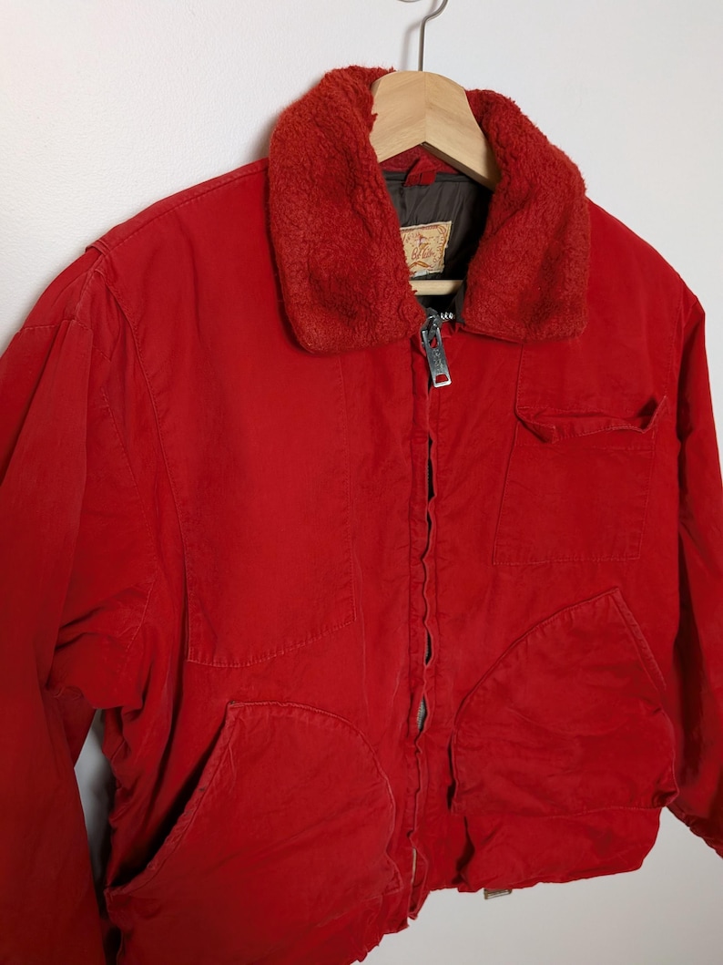 May include: A vibrant red jacket featuring a plush, reddish-brown collar. The jacket has a zip closure, two large front pockets, and a smaller chest pocket. The jacket is suspended on a wooden hanger.