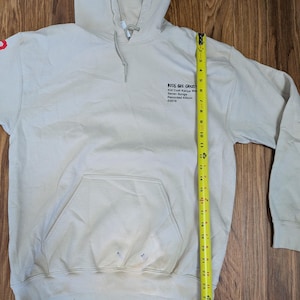 May include: A beige hooded sweatshirt with a front kangaroo pocket and a yellow measuring tape showing a length of 25 inches. The chest features black text reading KIDS SEE GHOSTS, Kid Cudi Kanye West, Seven Songs, Recorded Album, &copy;2018.