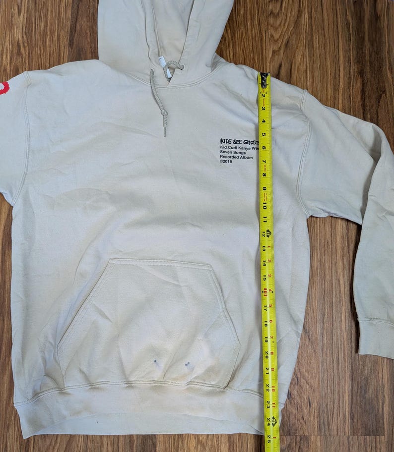 May include: A beige hooded sweatshirt with a front kangaroo pocket and a yellow measuring tape showing a length of 25 inches. The chest features black text reading KIDS SEE GHOSTS, Kid Cudi Kanye West, Seven Songs, Recorded Album, &copy;2018.