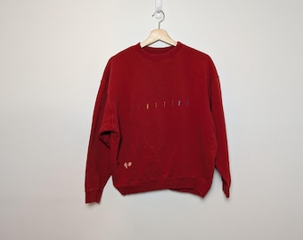 May include: A red sweatshirt with the word "SENTITIE" embroidered in colourful thread across the chest. The sweatshirt is hanging on a wooden hanger against a white wall. The garment has long sleeves and a crew neck.