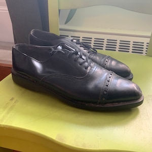 Może przedstawiać: Para czarnych skórzanych butów sznurowanych z noskiem typu captoe. Buty mają lekko zaokrąglony nosek i grubą podeszwę.