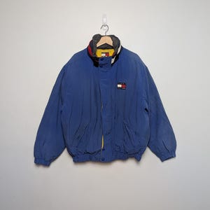 May include: A blue jacket with a high collar and a small logo patch. The jacket has a yellow lining and a red, white, and blue color scheme on the collar. It has two front pockets and a zipper closure.