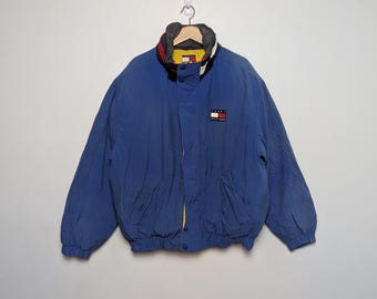 May include: A blue jacket with a high collar and a small logo patch. The jacket has a yellow lining and a red, white, and blue color scheme on the collar. It has two front pockets and a zipper closure.
