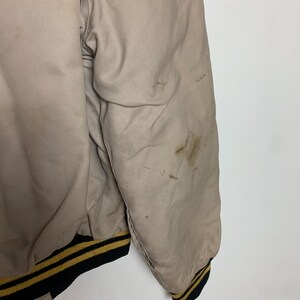 May include: A beige bomber jacket with black and gold striped ribbing at the cuffs and hem. The jacket has some light stains.