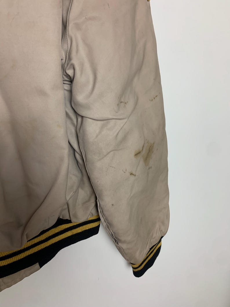 May include: A beige bomber jacket with black and gold striped ribbing at the cuffs and hem. The jacket has some light stains.