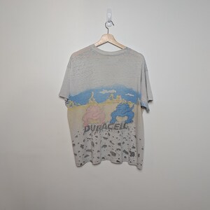 May include: A heather gray t-shirt with a colorful graphic design. The design features a landscape with blue and yellow hues, and two cartoon figures in pink and blue. The word "DURACELL" is printed below the figures.