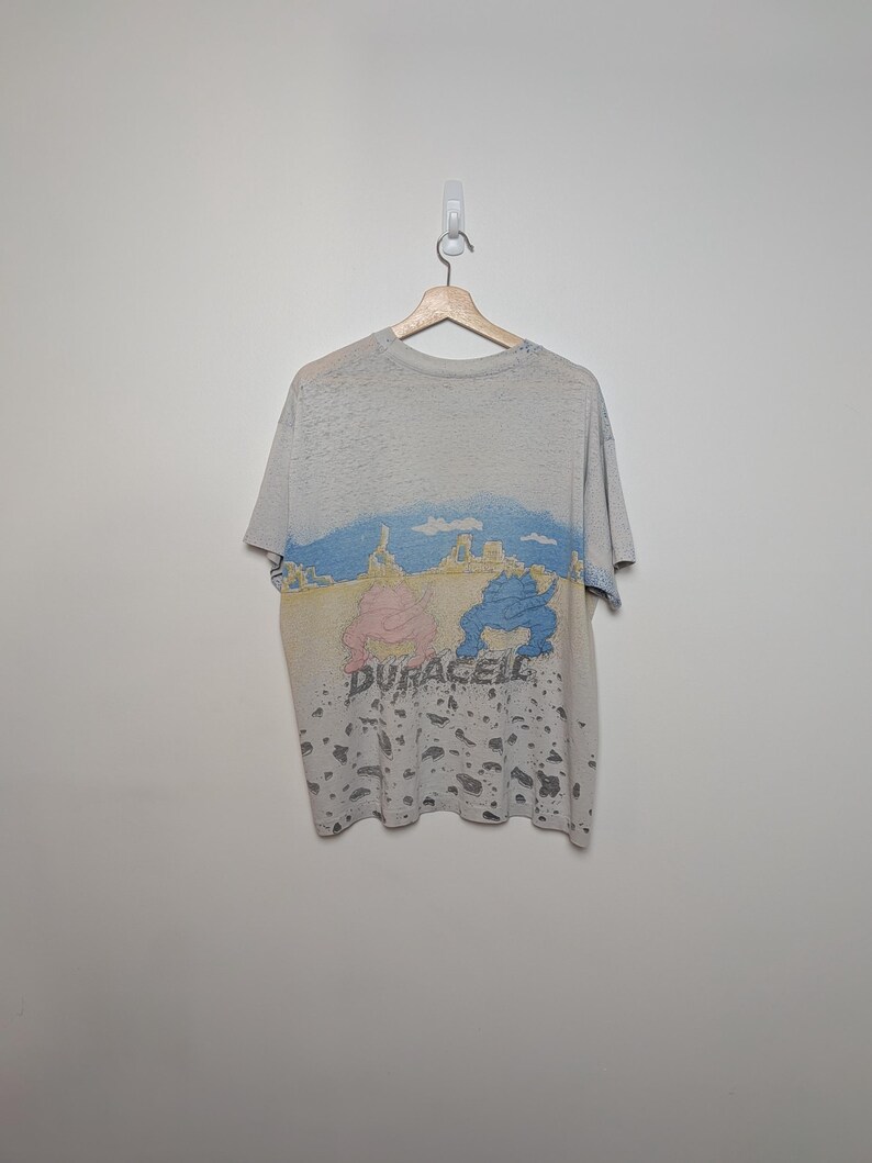 May include: A heather gray t-shirt with a colorful graphic design. The design features a landscape with blue and yellow hues, and two cartoon figures in pink and blue. The word "DURACELL" is printed below the figures.