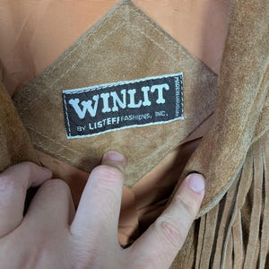 May include: A tan suede jacket featuring long fringe detailing and a diamond-shaped patch on the interior lining. The patch displays a black label with white text reading WINLIT BY LISTEFF FASHIONS, INC. and MADE IN URUGUAY in small print.