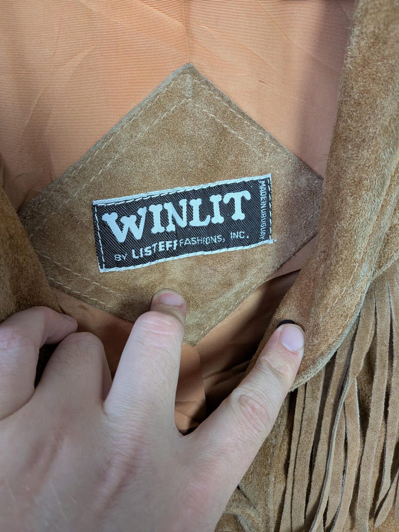 May include: A tan suede jacket featuring long fringe detailing and a diamond-shaped patch on the interior lining. The patch displays a black label with white text reading WINLIT BY LISTEFF FASHIONS, INC. and MADE IN URUGUAY in small print.