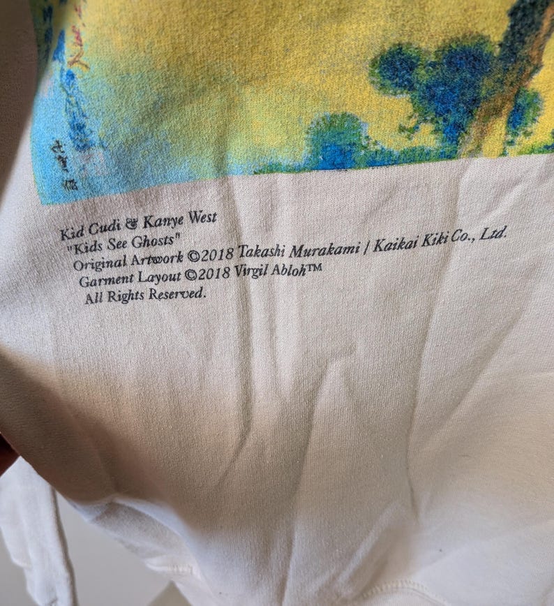 May include: A cream-colored sweatshirt featuring a vibrant yellow, blue, and green graphic print. Text reads: Kid Cudi & Kanye West "Kids See Ghosts" Original Artwork &copy;2018 Takashi Murakami / Kaikai Kiki Co., Ltd. Garment Layout &copy;2018 Virgil Abloh&trade;.
