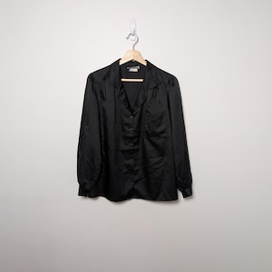 May include: A black button-down shirt with long sleeves, a collar, and a chest pocket. The shirt is hanging on a wooden hanger against a white wall. The fabric appears to be a smooth, possibly satin-like material.