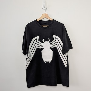 May include: A black short-sleeve t-shirt featuring a large, white, stylized spider graphic printed on the front. The design consists of a central body shape with long, angular legs extending outward toward the sides of the garment.