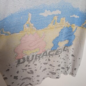 May include: A light gray t-shirt featuring a colorful graphic of two dinosaurs in a desert landscape with the word "DURACEL" printed below. The dinosaurs are pink and blue, with a yellow and blue sky.