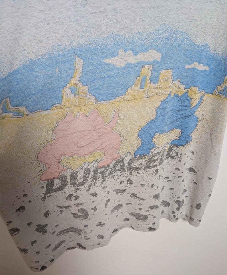 May include: A light gray t-shirt featuring a colorful graphic of two dinosaurs in a desert landscape with the word "DURACEL" printed below. The dinosaurs are pink and blue, with a yellow and blue sky.